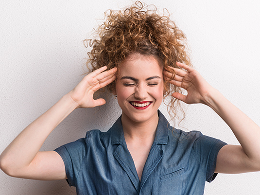 A woman with curly hair is smiling and holding her hands to the side of her head.